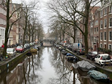 Amsterdam, Hollanda - 15 Ocak 2025: Amsterdam Kanalı serin bir kış gününde sakin bir su yolu boyunca parkedilmiş tekneler ve çıplak ağaçlar sergilenen bulutlu gökyüzünü yansıtır.