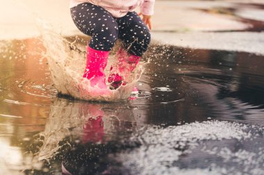 Feet of child in pink rubber boots jumping and splashing over puddle after rain. clipart
