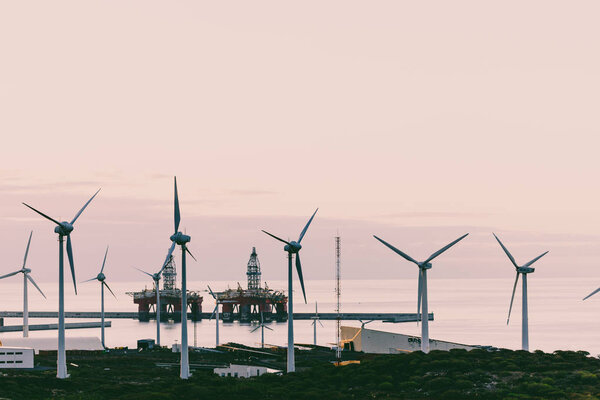 Group of windmills and solar panels for electric power production and oil rigs on coast.