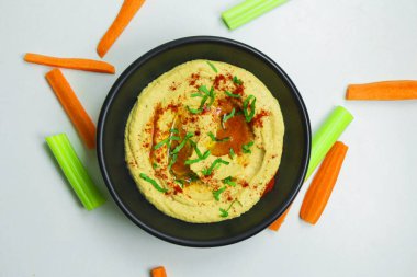 Zeytin yağı, kırmızı biber ve beyaz bir arka plan üzerinde siyah bir kase taze sebze ile ev yapımı humus