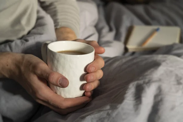 Hygge, kahve fincanı tutan bir adamla lagom yaşam tarzı imajı. Minimalist gri keten levha ile kaplı açık defter ve yatakta bir kalem ile arka plan. Rahat yaşam tarzı.
