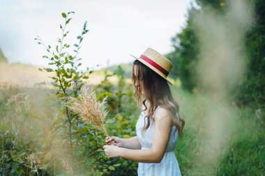 Hasır şapkalı gülen kız çiçek topluyor. Uzun otların arasında, yazlık elbiseli bir kız. Bir buket kurumuş çiçekle yeşil bir tarlada bir kadın.