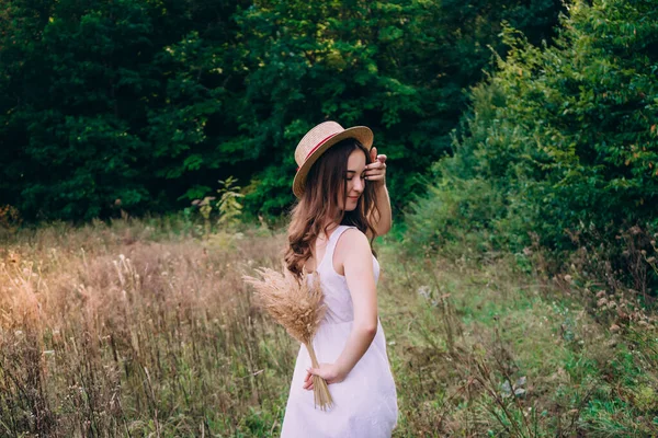 Beautiful girl with curly hair in a hat. Brunette girl walking in a field of flowers. Young woman in summer with a bouquet of dried flowers