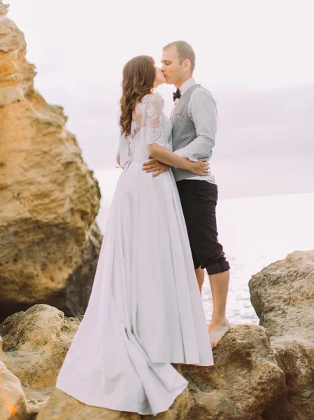 La foto al aire libre de la pareja recién casada besándose mientras está de pie en el acantilado en el fondo del mar . — Foto de Stock