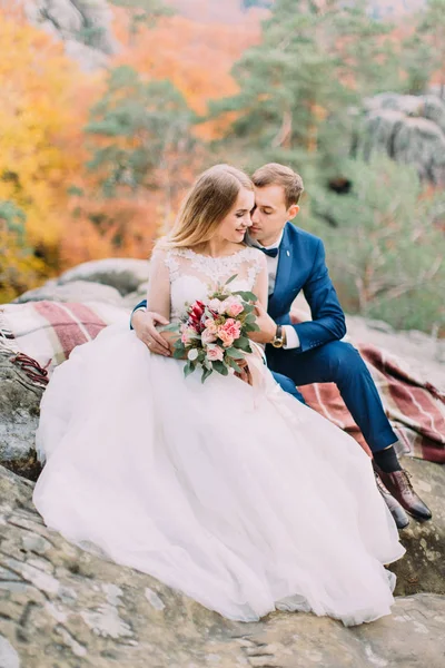 Foto completa de la pareja de recién casados abrazados sentados en las rocas y mirando a un lado . —  Fotos de Stock