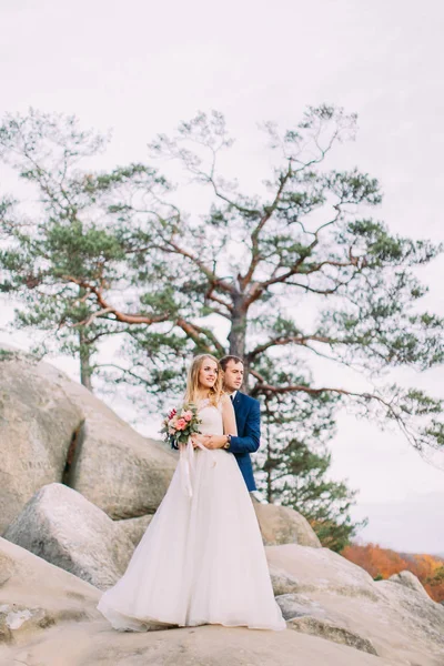 Full-length photo of the groom hugging the bride back while standing on the rock. — Stock Photo, Image