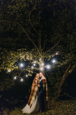 Garland lambalar ile dekore edilmiş ağaç aşağıda örme şal sarılı sarılma yeni evli çiftin tam uzunlukta fotoğraf.