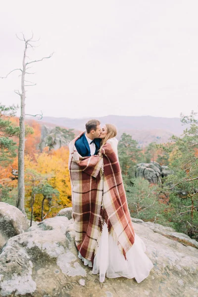 Full-length foto van het kussen jonggehuwde paar verpakt in de gebreide plaid. Locatie van de bergen. — Stockfoto