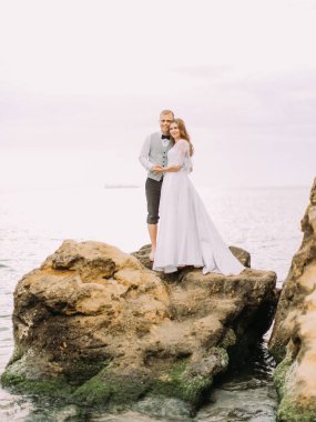 Gülümseyen yeni evliler günbatımı sırasında deniz arasında Uçurumdaki duruyorsun.