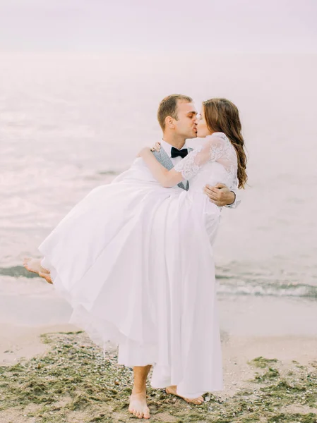 El novio sostiene a la novia en sus brazos mientras la besa. La composición del mar . —  Fotos de Stock