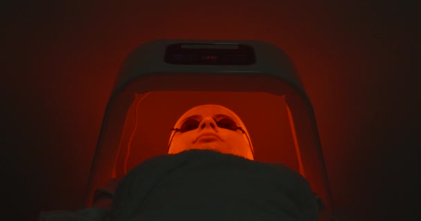 Terapia de luz led para la cara de la mujer joven con máscara y gafas que yacen en cabina especial en el salón de spa. Cambiando de color. Imágenes de 4k — Vídeos de Stock