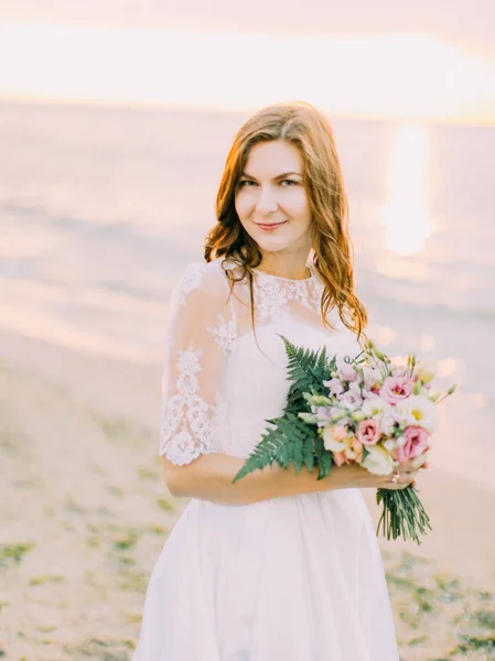 O lindo retrato da noiva sorridente segurando o buquê de casamento no fundo do pôr do sol . — Fotografia de Stock