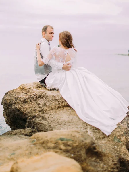 Widok z tyłu nowożeńcy, siedząc na klifie i patrząc na siebie na tle morza. — Zdjęcie stockowe