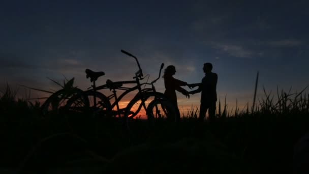 Paseo romántico con bicicletas. Siluetas de la sensible pareja amorosa bailando en el campo sobre el cielo rojo y gris durante la puesta del sol . — Vídeos de Stock