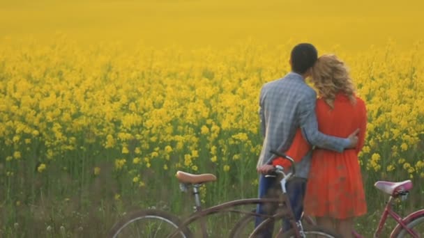 Vista trasera de cerca de la atractiva pareja de amantes abrazándose tiernamente y besándose mientras disfruta de la naturaleza del encantador campo cubierto de flores amarillas florecientes. Caminar con bicicletas . — Vídeo de stock