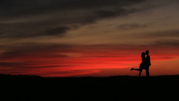 Paseo romántico de la adorable pareja besándose tiernamente sobre el cielo rojo y gris durante la puesta del sol. Vista completa de las siluetas . — Vídeo de stock
