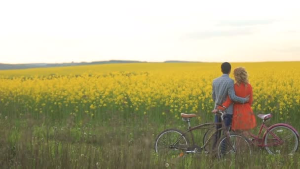 Vista trasera de la pareja de amantes con bicicletas abrazándose tiernamente mientras disfruta de la paliza del campo cubierto de flores amarillas florecientes . — Vídeos de Stock