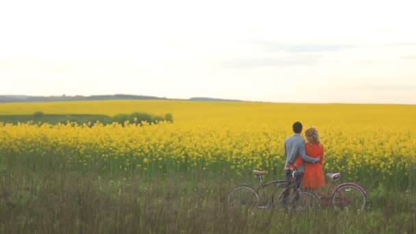 Bedårande älskande par med cyklar är mjukt kramas och njuter av den vackra vackra naturen i fältet täckt med gula blommor. Bakifrån. Inget ansikte. — Stockvideo