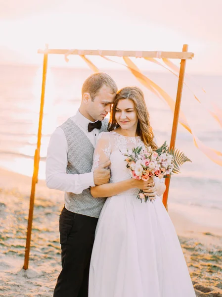 Preciosa composición de los recién casados abrazándose en la playa durante la puesta del sol . — Foto de Stock