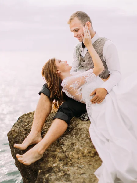 Il ritratto laterale dello sposo seduto sulla scogliera mentre la sposa è sdraiata sulle ginocchia e accarezzando il suo viso sullo sfondo del mare . — Foto Stock