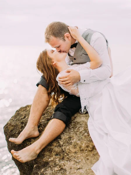 Das Outdoor-Porträt des küssenden Brautpaares. der Bräutigam sitzt auf der Klippe, während die Braut auf seinem Schoß am Hintergrund des Meeres liegt. — Stockfoto