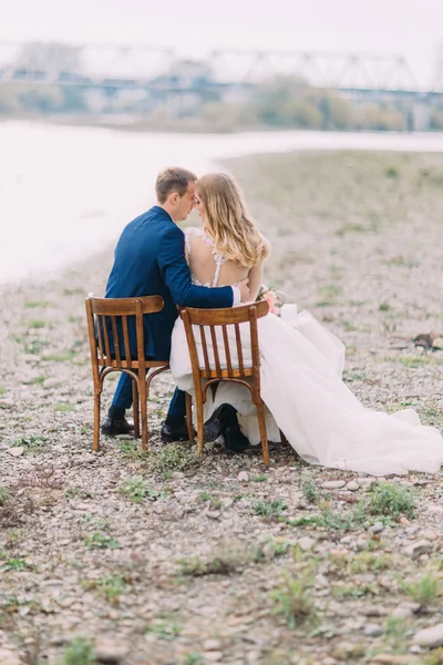 Hellång tillbaka beskåda av de krama nygifta medan du sitter på stolarna vid kusten. — Stockfoto