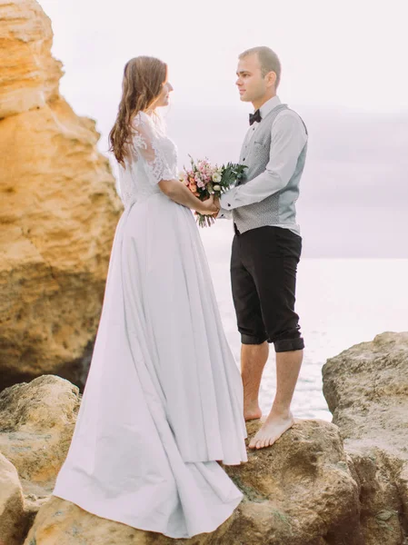 A teljes hosszúságú fotó a boldog ifjú házasok, kézen állva a sziklafalon a háttérben a tenger. — Stock Fotó