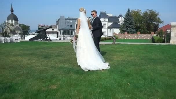Bakifrån av unga attraktiva par av nygifta hålla händerna medan du går längs trädgården nära slottet. Brudgummen ömt kramar och kysser bruden i huvudet. — Stockvideo