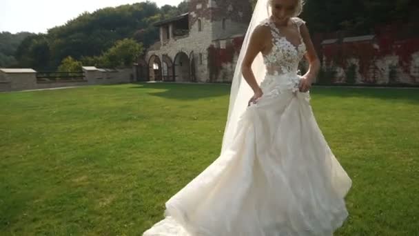 Rear full-length view of the young charming blonde bride in the beautiful long dress running along the blooming garden. — Stock Video