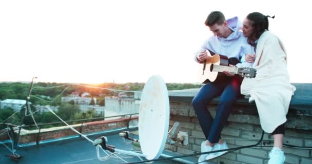 Romantisk atmosfär, vackra lyckliga paret sitter o nOLE taket. Den stiliga killen spelar gitarr och härlig tjej insvept i rutigt lyssnar på med leende. — Stockvideo