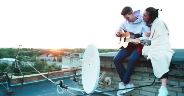 Romantische Atmosphäre, schönes glückliches Paar sitzt auf dem Dach. der gutaussehende Kerl spielt Gitarre und das hübsche, in Plaid gehüllte Mädchen hört mit dem Lächeln zu. — Stockvideo