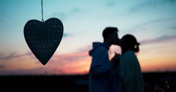 Close-up view of the hanging metal heart at the blurred background of the sensitive loving couple tenderly kissing over the colourful sky during the sunset. — Stock Video