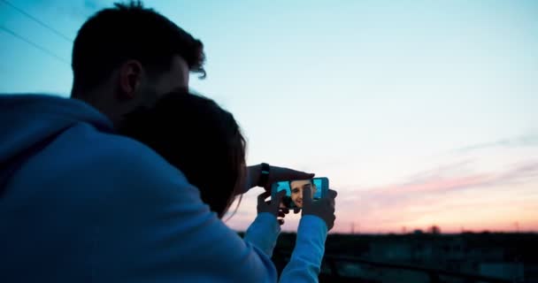 Achteraanzicht van het gelukkig lachend verliefde paar kijken naar foto's op de mobiele telefoon en genieten van de zonsondergang op het dak. — Stockvideo