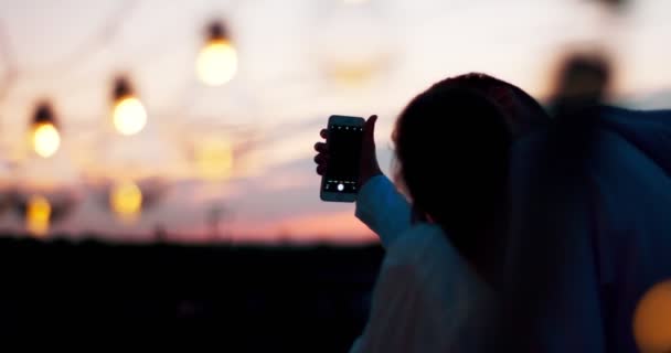 Achteraanzicht van het gelukkige lachende paar van twee geliefden selfies te nemen tijdens de zonsondergang op het dak is voorzien van tekenreeks pf lampen. 4 k-beeldmateriaal. — Stockvideo