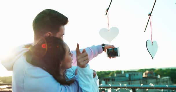 Vue arrière du couple affectif heureux embrassant et prenant selfies sur le toit décoré de coeurs. Ambiance romantique. Images 4k . — Video