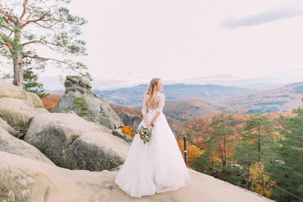 Achteraanzicht van de bruid de bruiloft boeket permanent op de rots en genieten van het uitzicht op de vergeelde bos. — Stockfoto
