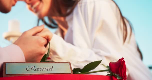 Composição romântica. Placa Reservado e vermelho rosa sobre a mesa no fundo borrado do casal no amor ternamente de mãos dadas, esfregando narizes e beijando . — Vídeo de Stock