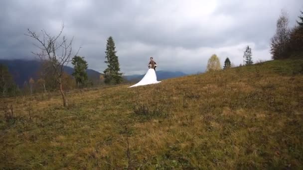Full-length view at the stylish couple of newlyweds in love softly hugging on the top of the mountains in autumn during the cloudy day. — Stock Video