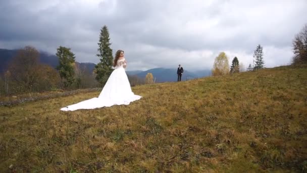 スタイリッシュな新郎は秋の山の上にファッショナブルなドレスで美しい魅力的な新妻に来ています。. — ストック動画