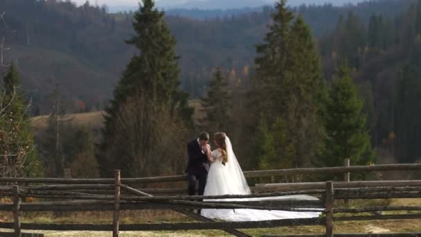 Hochzeit in den Bergen. Der attraktive Bräutigam hält und küsst seine wunderbare Braut während des Dorfspaziergangs. Ansicht in voller Länge. — Stockvideo