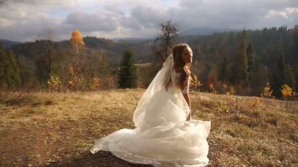 Encantadora noiva engraçada em vestido lindo está se divertindo enquanto dança e pulando com o véu nas montanhas douradas . — Vídeo de Stock