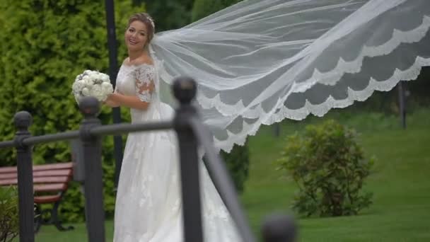 Allurning sposa sorridente in abito splendido sta tenendo il bouquet di nozze di peonie bianche. Il vento sta muovendo il suo lungo velo . — Video Stock