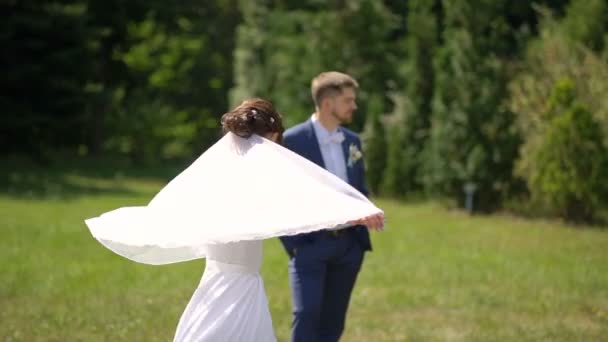 La splendida sposa bruna in bellissimo abito da sposa sta girando, giocando e chiudendo il velo con il velo sullo sfondo sfocato dello sposo . — Video Stock