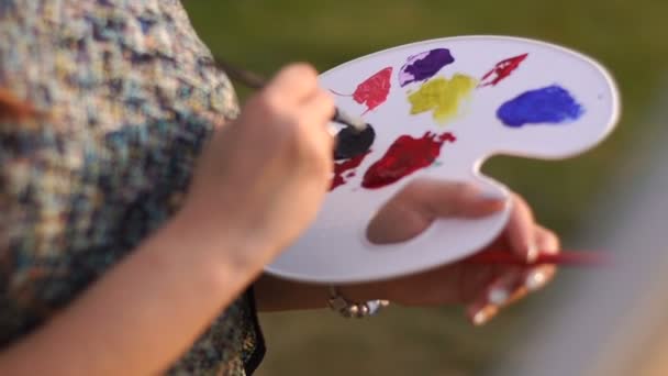 Unknown woman is moxing colors on the artistic palette and plainting the picture in the sunny field. — Stock Video