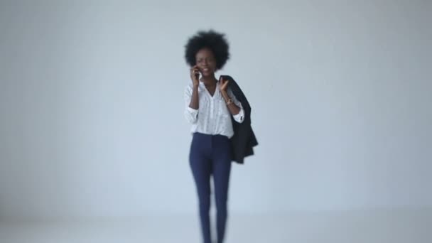 Vista a figura intera della giovane bella donna africana in tessuto casual sui tacchi alti sta parlando sul telefono cellulare mentre cammina lungo lo studio bianco . — Video Stock