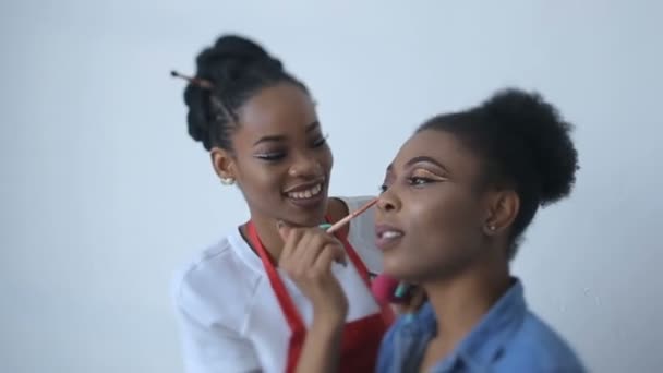 Charmiga leende make-up artist tillämpar make-up på framsidan underbara afrikanska modell. Skönhet-konceptet. — Stockvideo