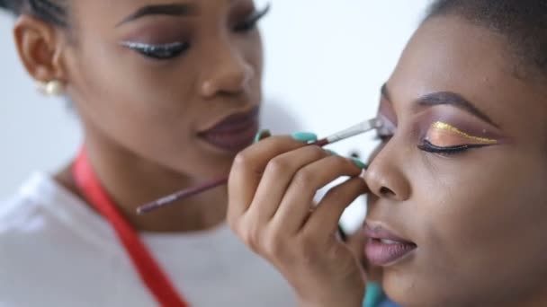 Maquillador profesional que aplica maquillaje con el pincel en los párpados del encantador modelo africano elegante . — Vídeos de Stock