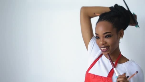 Retrato del joven y adorable maquillador africano con una bonita sonrisa sosteniendo el conjunto de pinceles y paletas sobre fondo blanco . — Vídeos de Stock