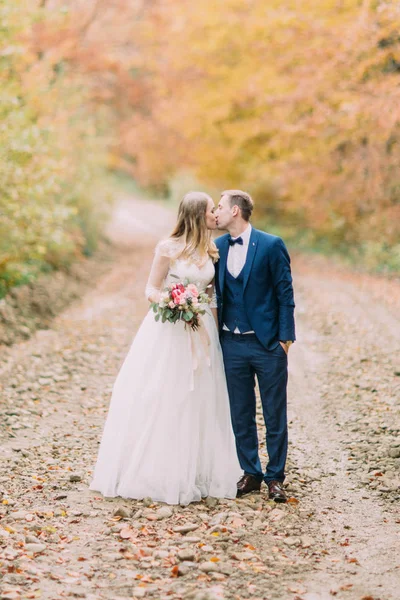 Visão completa dos recém-casados beijando na estrada perto da madeira do outono . — Fotografia de Stock