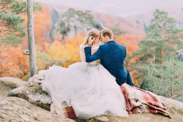 O noivo sorridente está tocando a cabeça da noiva feliz enquanto está sentado na rocha na floresta amarelada . — Fotografia de Stock